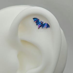 May include: A small, iridescent blue and purple bat charm for an ear piercing. The charm is on a white ear model.