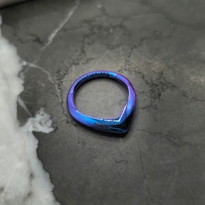 May include: A blue and purple iridescent seamless ring made of surgical steel. The ring is shaped like a teardrop and has a smooth, polished finish.