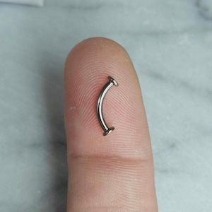 May include: A curved, silver-colored eyebrow piercing. The jewelry has a slight arc with flat, circular ends. The piece is small and appears to be made of metal. The piercing is shown on a fingertip.
