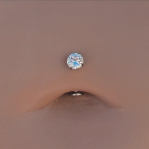 May include: A navel piercing jewelry piece featuring a single, round, clear faceted gemstone set in a silver-toned metal prong setting. The circular stone is positioned above the navel opening, with a small metal backing visible inside the navel.