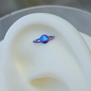 May include: A small, iridescent planet-shaped earring stud. The stud features a rounded, blue-purple sphere with a ring around it, and is set against a white background. The earring is a unique piece of jewelry.