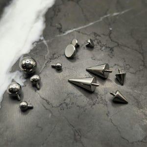 May include: A collection of silver-toned metal body jewelry attachments featuring various shapes including smooth spheres, flat circular discs, and pointed cones. Each piece includes a small threaded screw post for secure fastening.