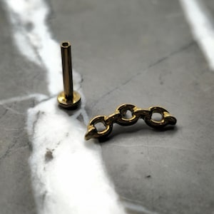 May include: A gold-colored body piercing stud and a small chain link charm. The stud has a flat base and a cylindrical post. The chain link charm has three connected links.