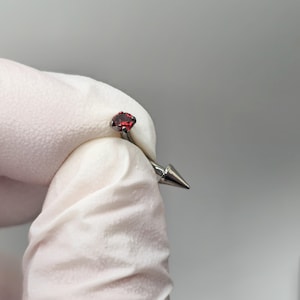 May include: A curved barbell body jewelry piece featuring a round, faceted red gemstone set in a silver-toned metal prong setting on one end and a smooth, pointed silver-toned metal cone on the opposite end of the curved metal bar.