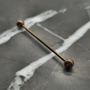 May include: A gold-colored industrial barbell earring. The earring features a straight bar with a ball at each end. The jewelry is displayed on a gray and white marbled surface.