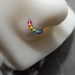 May include: A rainbow-colored nose ring featuring small, round beads in a spectrum of colors: red, orange, yellow, green, blue, and purple. The beads are strung together on a thin, silver-colored wire, forming a curved shape.