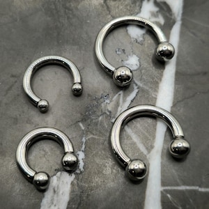 May include: Four silver horseshoe-shaped body jewelry pieces with a polished finish. Each piece has a rounded ball at each end. The pieces vary in size, with the smallest at the top left and the largest at the bottom right.