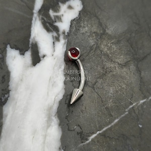 May include: A silver curved barbell with a red gem on the top and a pointed end on the bottom. The barbell is on a gray and white marble surface.