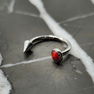 May include: A silver-colored, curved barbell-style piercing ring. One end features a conical spike, while the other has a round, red gemstone. The jewelry is set against a marbled background.