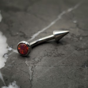 May include: A silver body piercing with a red opal gemstone and a pointed end.