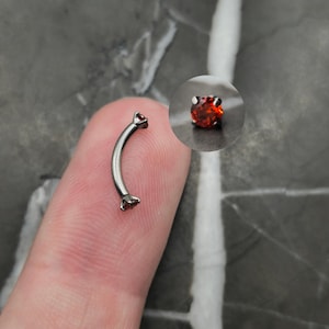 May include: Curved eyebrow piercing jewelry with a silver-tone metal bar and two red gemstone ends. The jewelry is shown close-up, with a small, detailed inset image of a single gemstone.