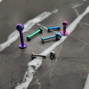 May include: A collection of six metal body piercing studs in various colors including purple, green, blue, and silver. Each stud features a flat circular base, a straight post, and a small spherical top. One silver stud is disassembled into two parts.