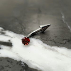 May include: A silver body piercing with a red gem and a pointed end. The piercing is curved and designed for a belly button piercing.