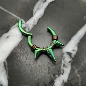 May include: A green septum ring with three conical spikes and brown wire accents. The curved bar is open, and the spikes are evenly spaced. The jewelry is set against a marbled gray background.