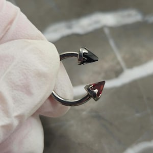 May include: A circular horseshoe-shaped body jewelry piece featuring a metallic silver-toned band. One end is capped with a pointed black stone, while the opposite end is capped with a pointed deep red stone, both set in metallic silver-toned prongs.
