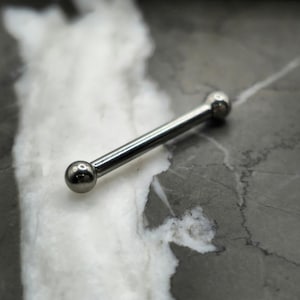 May include: A silver barbell-style piercing jewelry with a straight bar and two round ball ends. The jewelry is made of a shiny, metallic material. The piercing is shown against a marbled gray and white background.