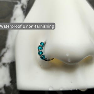 May include: A silver-toned nose ring hoop featuring four small, round, translucent teal beads secured by wire wrapping. The text 'Waterproof & non-tarnishing' is visible on the image. The jewelry is displayed on a white nose model.