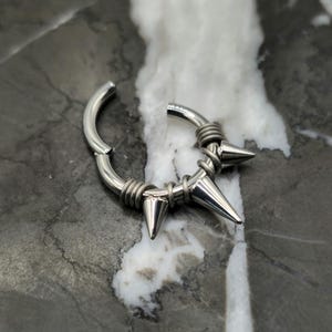 May include: A silver-colored, hinged hoop earring with a spiked design. The earring features three conical spikes and is wrapped with wire detailing. The jewelry is shown against a marbled background.