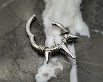 May include: A silver-colored, hinged hoop earring with a spiked design. The earring features three conical spikes and is wrapped with wire detailing. The jewelry is shown against a marbled background.