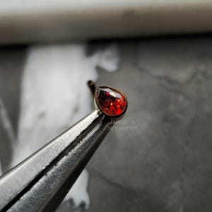 May include: A close-up of a red teardrop-shaped gemstone nose ring. The ring is held in a pair of metal tweezers. The text "Embers of Moonlight" is visible in the image.