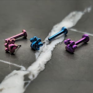 May include: Four pieces of body jewelry with paw print designs. The jewelry is in shades of pink, blue, and purple. The pieces are on a dark, marbled surface.