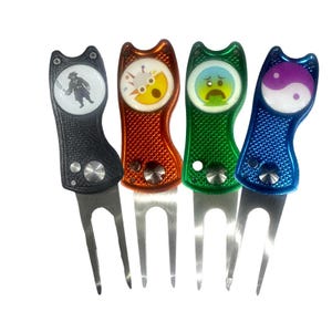 Peut inclure: Quatre outils de réparation de divots de golf en noir, rouge, vert et bleu. Chaque outil a un dessus en forme de chat avec un graphique différent : un pirate, un soleil, un visage triste et un symbole yin-yang. Les outils ont une fourche en métal.