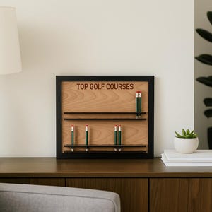 May include: A wooden display with the text "TOP GOLF COURSES" and several pencils. The display is framed in black and sits on a dark wood surface. A white lamp and a plant are also visible.