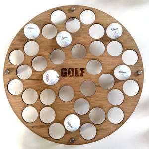 May include: A circular wooden golf ball display featuring the word "GOLF". The display has numerous circular recesses, some containing golf balls. The golf balls are white with black text and the number "3". Metal fixings secure the display.