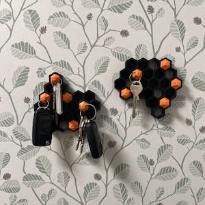 以下が含まれることがあります： オレンジ色のアクセントが付いた黒いハニカム型のキーホルダーが2つ、花柄の壁に取り付けられています。キーホルダーにキーが掛けられています。