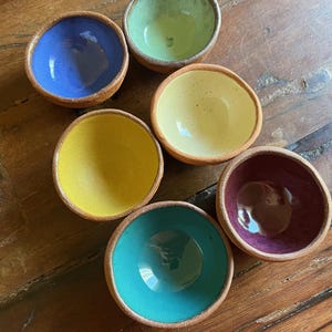 May include: Six small, handmade ceramic bowls with a natural clay exterior and vibrantly colored interiors. The bowls feature a variety of colors, including blue, green, yellow, and purple. The bowls are arranged on a wooden surface.