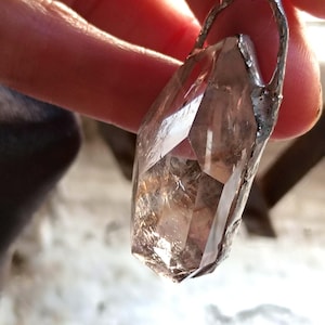 May include: A clear, faceted crystal pendant with a silver-coloured metal setting. The crystal has a natural, smoky appearance with internal inclusions. The pendant is suspended from a delicate chain, showcasing a unique, earthy aesthetic.