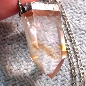 May include: A clear quartz crystal pendant with a silver-colored metal cap and chain. The crystal is a light amber color with internal inclusions and facets. The chain is a delicate, linked design.