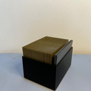 Könnte beinhalten: Eine schwarze Box mit einem Stapel brauner Karten darin. Die Box ist aus robustem Material gefertigt und hat ein elegantes Design.