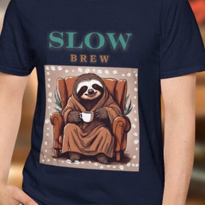 以下が含まれることがあります： ネイビーブルーのTシャツ。 ターコイズ色の「SLOW BREW」の文字と、コーヒーカップを持ったローブ姿のナマケモノのグラフィック。 ナマケモノは茶色の肘掛け椅子に座っています。 このシャツはカジュアルウェアです。