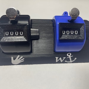 May include: A black and blue mechanical counter with a hand and anchor design. Each counter displays "0000" and has a silver lever. The base is black with a textured surface.