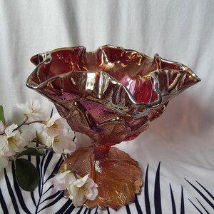 May include: A decorative, iridescent glass bowl with a ruffled rim and a pedestal base. The bowl transitions from red to gold hues, with a silver-colored rim. The base is shaped like a large leaf. The bowl is a decorative item.