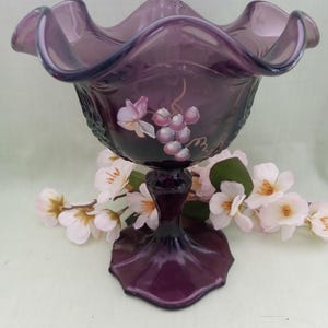 May include: A purple glass candy dish with a ruffled rim and a painted grape design. The dish is on a pedestal base.