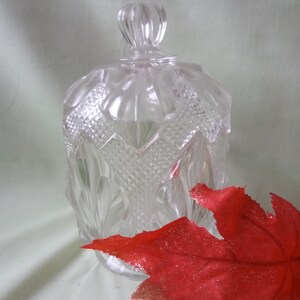 May include: A clear glass jar with a lid, featuring a diamond pattern design. The jar is sitting on a white surface with a red leaf in the foreground.