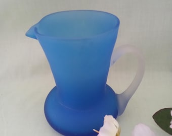 May include: A small, frosted blue glass pitcher with a rounded body and a handle. The pitcher is perfect for serving small amounts of liquid.