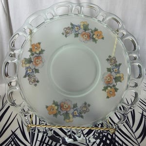 May include: A clear glass plate with a scalloped edge and floral design. The plate features clusters of orange, yellow, and blue flowers. The plate is displayed on a gold-colored metal stand, set against a black and white patterned background.