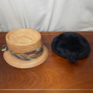May include: Two hats on a wooden surface. One is a straw boater hat with a plaid band in shades of green, brown, and tan. The other is a black, fuzzy hat with a small, attached detail. The hats are side by side.