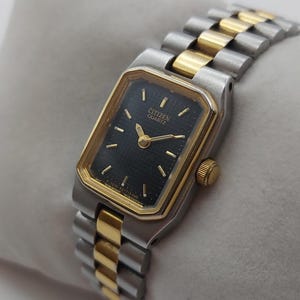 Könnte beinhalten: Eine Citizen Quartz-Uhr mit einem rechteckigen schwarzen Zifferblatt und goldfarbenen Akzenten. Die Uhr hat ein zweifarbiges silbernes und goldenes Armband. Die Uhr wird auf einem weißen Kissen präsentiert.