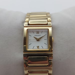 May include: Gold-toned Orient wristwatch with a rectangular white face and Roman numeral hour markers. The watch has a gold link band and is displayed on a white pillow. The watch face reads "Orient".