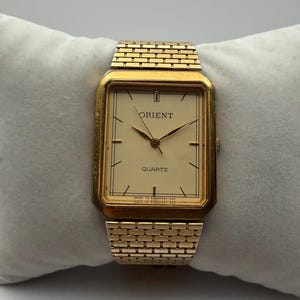 Puede incluir: Un reloj Orient Quartz rectangular dorado con esfera color crema y agujas doradas. El reloj tiene una correa dorada estilo ladrillo. El reloj se muestra sobre una almohada blanca.