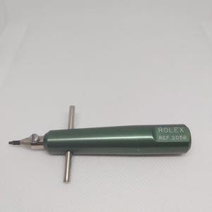 Puede incluir: Una herramienta de metal verde con un mango plateado y una punta puntiaguda. La herramienta está etiquetada con "ROLEX REF 2056".
