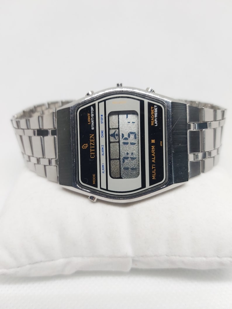 May include: A silver Citizen Multi Alarm III watch with a black face and a digital display. The watch has a silver metal strap and features buttons for light, start/stop, alarm, and lap/reset.