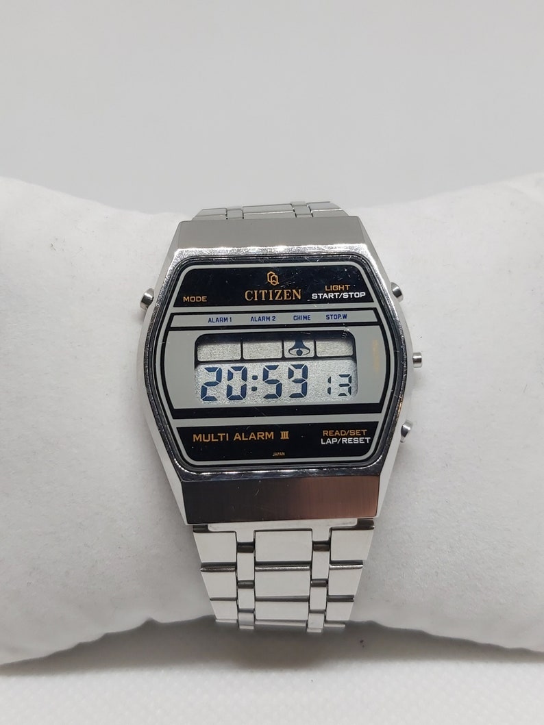 May include: Silver Citizen digital watch with a black face and a silver metal strap. The watch face displays the time, date, and has buttons for alarm, chime, stopwatch, and light.