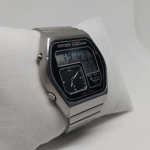 May include: A silver Citizen watch with a black face and a digital display. The watch has a chronograph, alarm, and chime functions. The watch face displays the time, date, and day of the week. The watch has a silver metal band.