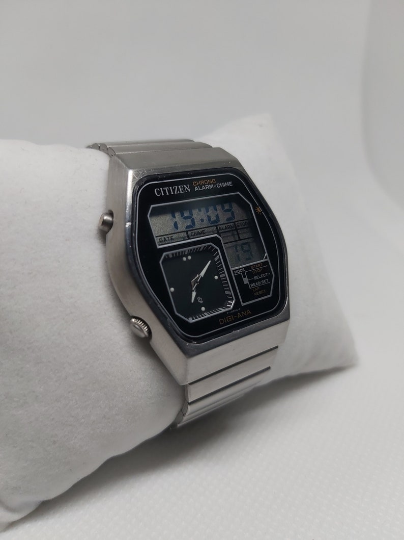 May include: A silver Citizen watch with a black face and a digital display. The watch has a chronograph, alarm, and chime functions. The watch face displays the time, date, and day of the week. The watch has a silver metal band.