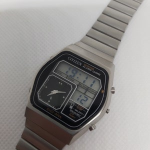 May include: A silver Citizen watch with a black face and a digital display. The watch has a chronograph, alarm, and chime functions. The watch face also has a small analog display with a black background and silver hands.